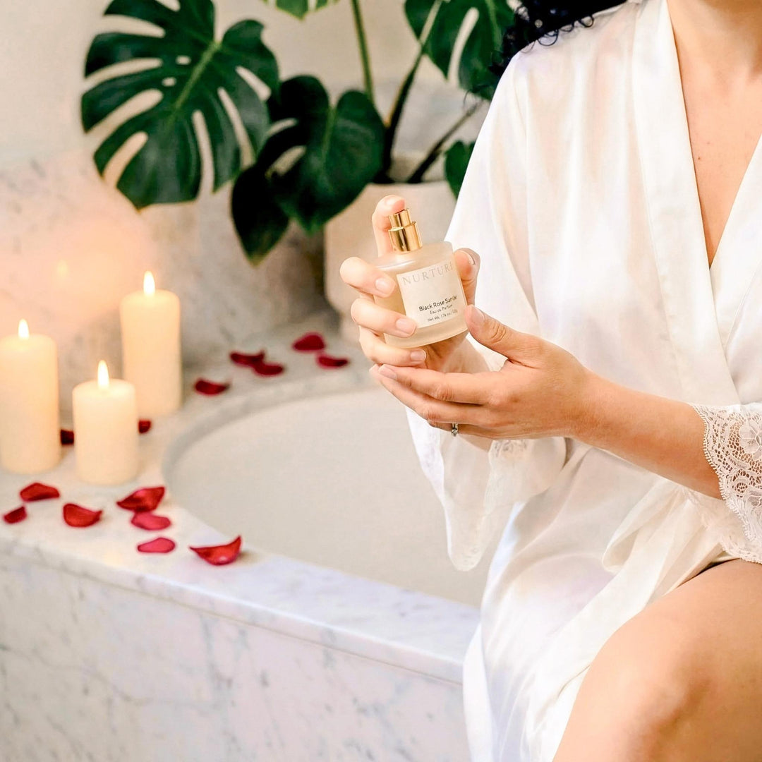 Person in a white robe holding a bottle of perfume with candles and rose petals in the background