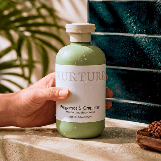 A hand is holding a bottle of Bergamot body wash on a shower shelf.