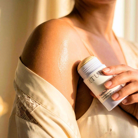 Person applying deodorant  to shoulder with a bottle labeled Patchoui Rose