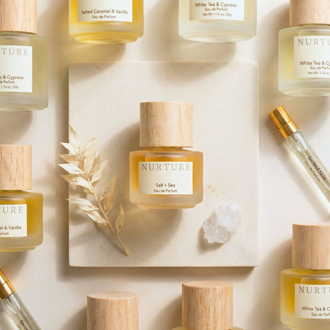 A styled flat-lay of NURTURE Eau de Parfum bottles with light wood caps, arranged on a cream background with a central square tile, dried grass, and a crystal.