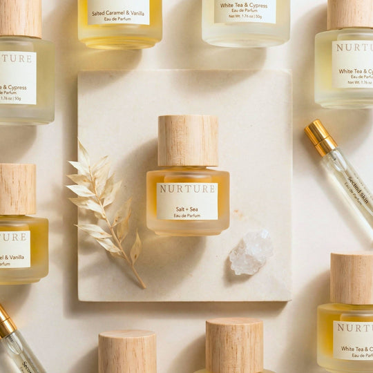 A styled flat-lay of NURTURE Eau de Parfum bottles with light wood caps, arranged on a cream background with a central square tile, dried grass, and a crystal.