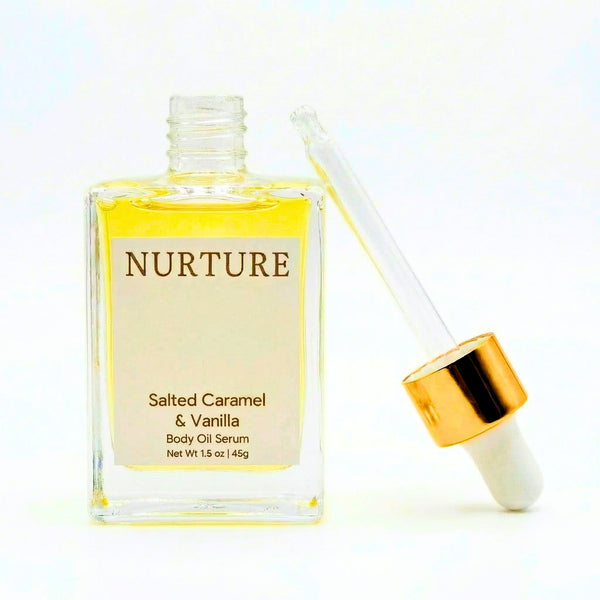 A clear glass bottle of Nurture Salted Caramel & Vanilla Body Oil Serum with a cream-colored label, shown next to its clear dropper with a shiny gold collar, on a white background.