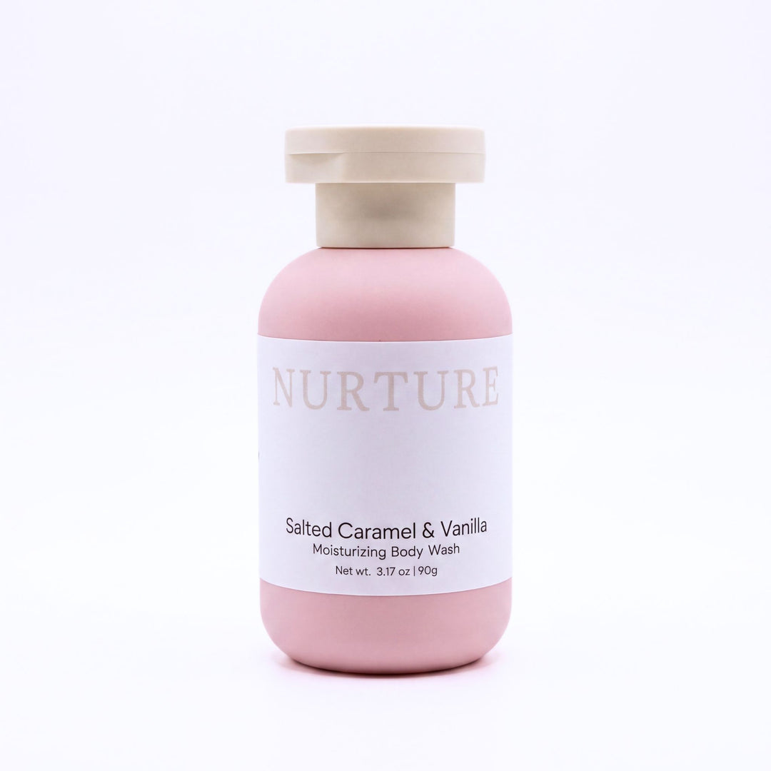 A bottle of NURTURE Salted Caramel & Vanilla Moisturizing Body Wash, featuring a matte pink bottle and a light beige cap, set against a clean white background.