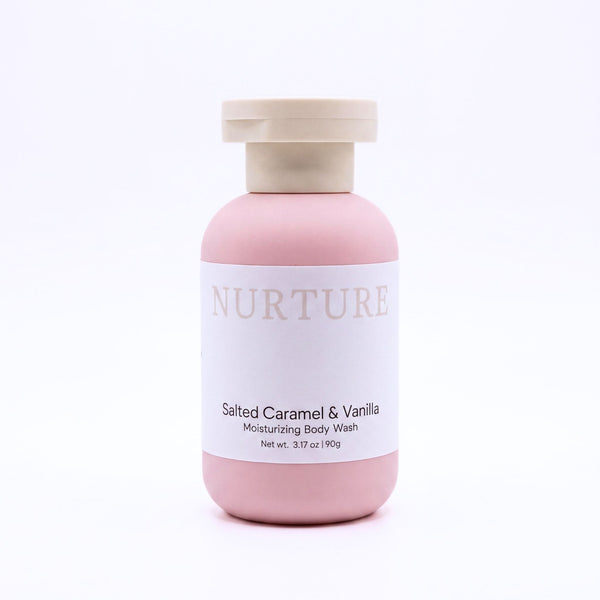A bottle of NURTURE Salted Caramel & Vanilla Moisturizing Body Wash, featuring a matte pink bottle and a light beige cap, set against a clean white background.