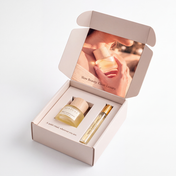 An open blush pink gift box on a white background containing a small amber bottle with a light wood cap and a slim gold-and-clear roller bottle. The inside lid of the box features a warm-toned printed photo with the text 'Slow Beauty, Clean Luxury.