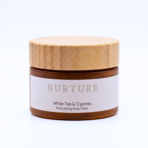 A jar of NURTURE White Tea & Cypress Moisturizing Body Polish with a light wood lid and a white label, set against a plain white background.