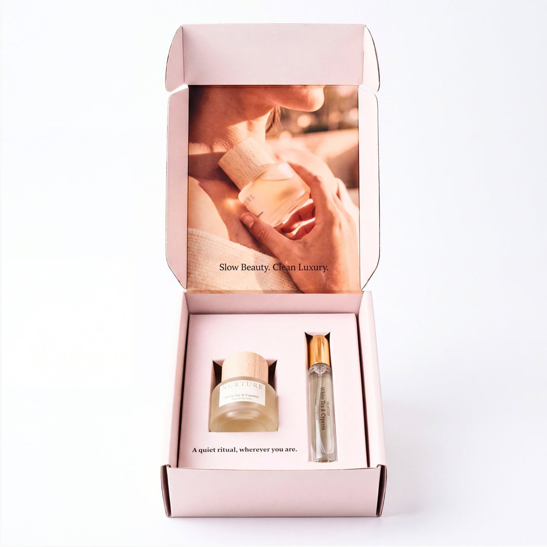 An open light pink gift box on a white background, containing a small frosted jar and a slim cylindrical bottle with a metallic cap. The inside lid features a warm-toned photo of hands holding a bottle and the text &