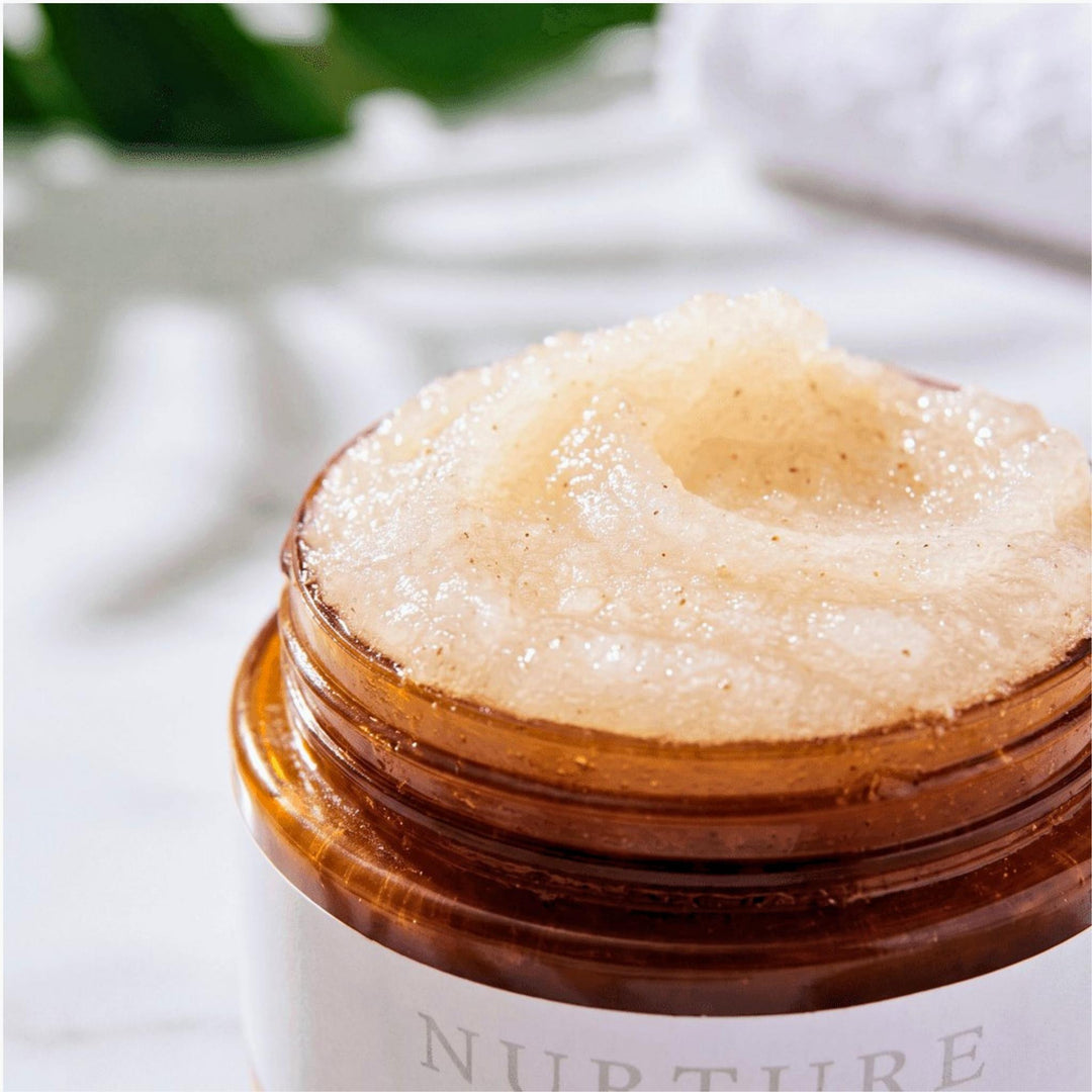 A close-up of a clear amber jar filled with a thick, pale beige textured Nurture Body Polish, with a white label partially visible at the bottom. The background is softly blurred with white and green tones.