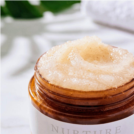 A close-up of a clear amber jar filled with a thick, pale beige textured Nurture Body Polish, with a white label partially visible at the bottom. The background is softly blurred with white and green tones.