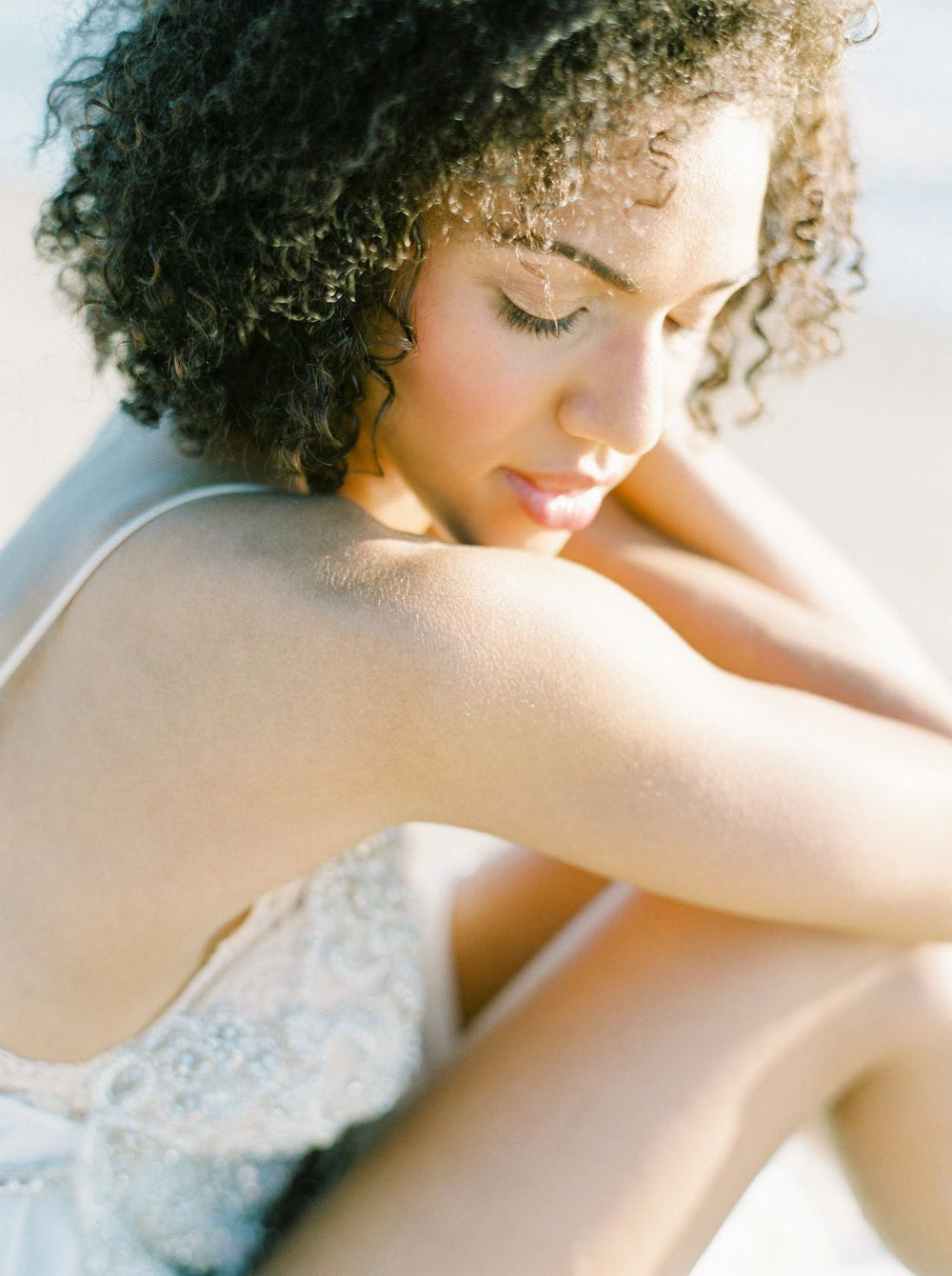Woman with curly hair wearing a white dress with lace details, sitting outdoors.
