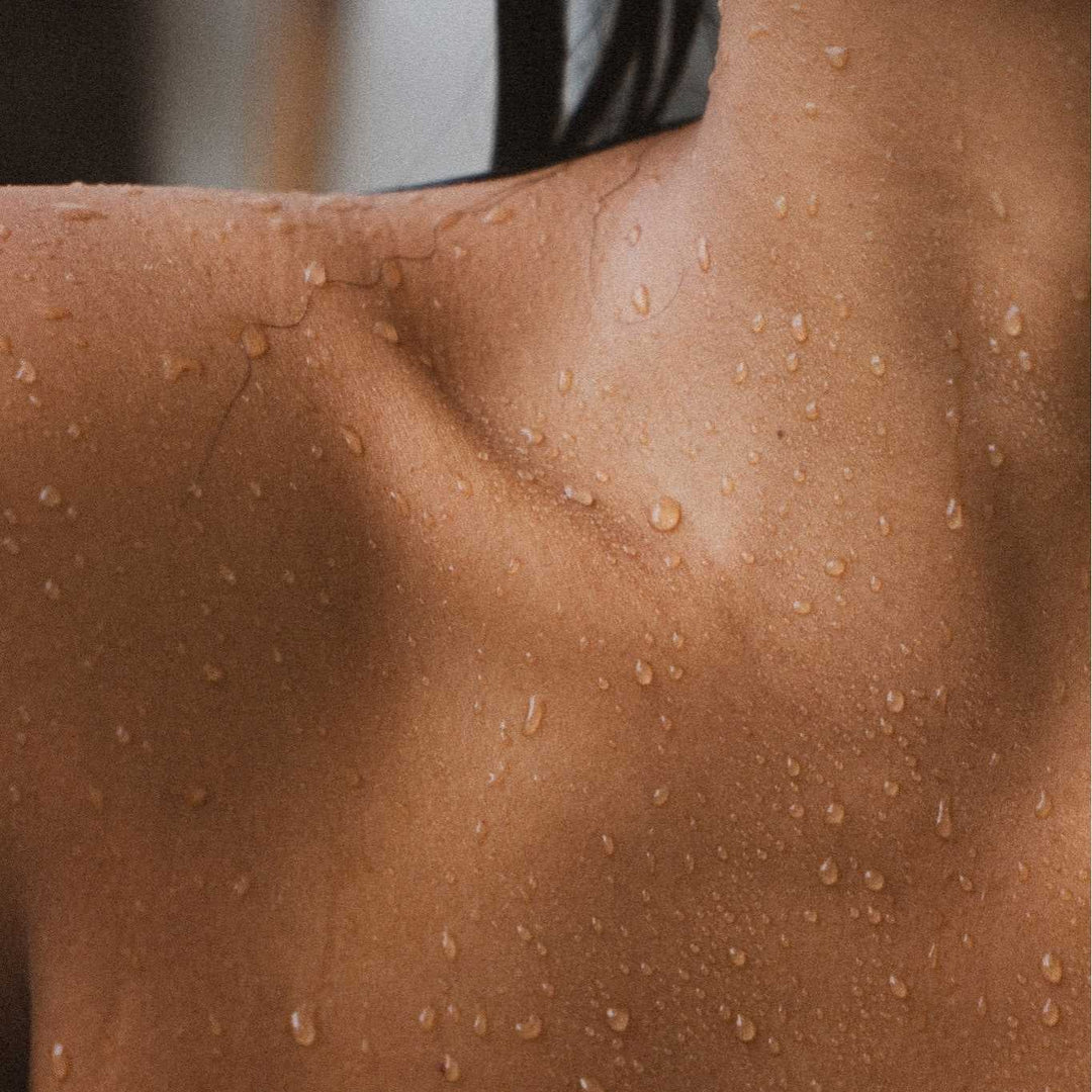 Close-up of a person's bare upper chest and shoulder covered in small water droplets, with a hand partially visible near the lower edge.